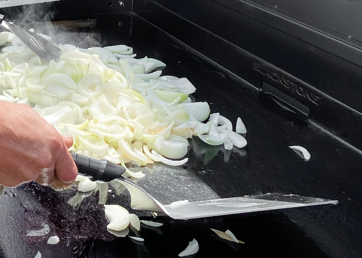 sliced onions being cooked on a large Blackstone griddle with metal spatulas