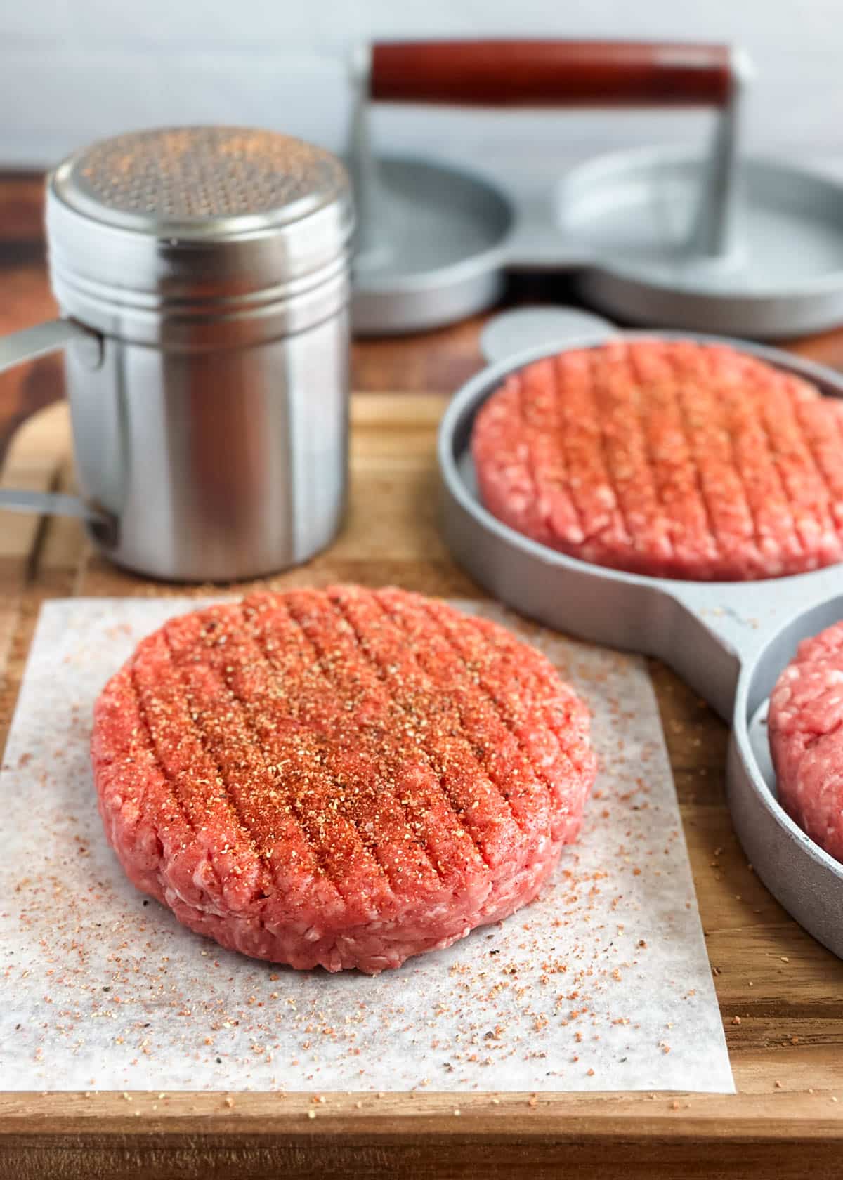 raw hamburger patty on parchment seasoned with burger seasoning, with a silver dredging shaker in the back