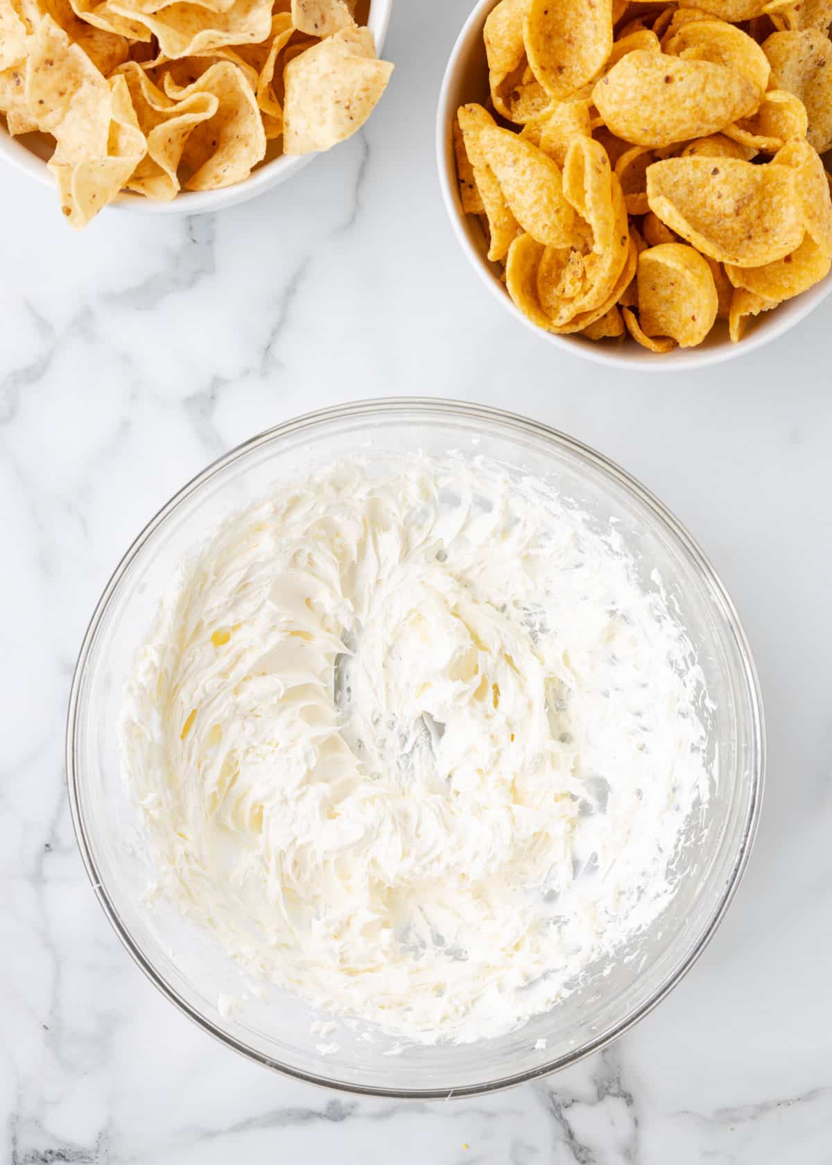 overhead of whipped cream cheese in a glass bowl on a marble board next to bowls of corn chips