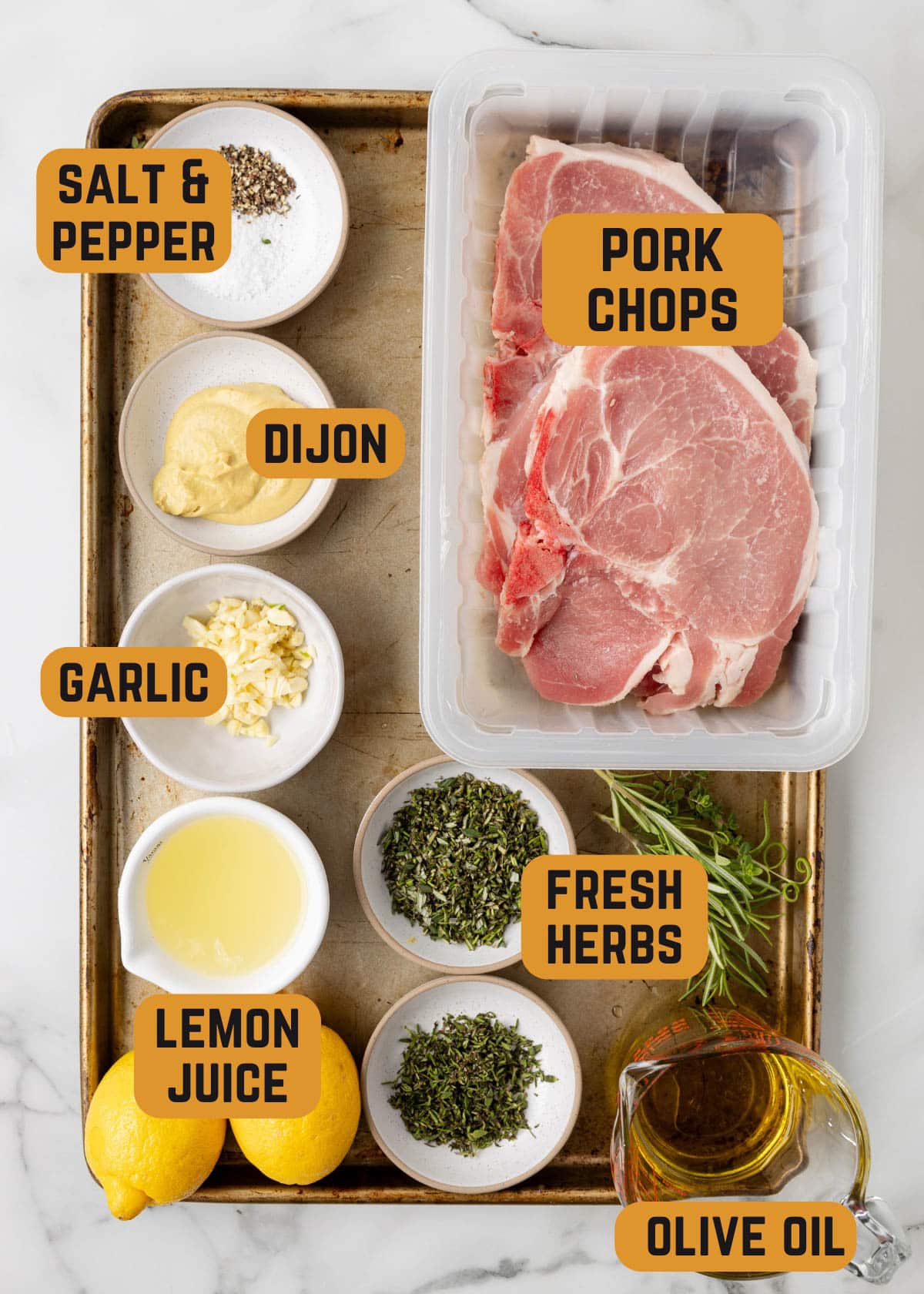 overhead photo of ingredients needed to make grilled lemon herb pork chops on a baking sheet. Text overlays read "salt & pepper, Dijon, garlic, lemon juice, pork chops, fresh herbs, olive oil"