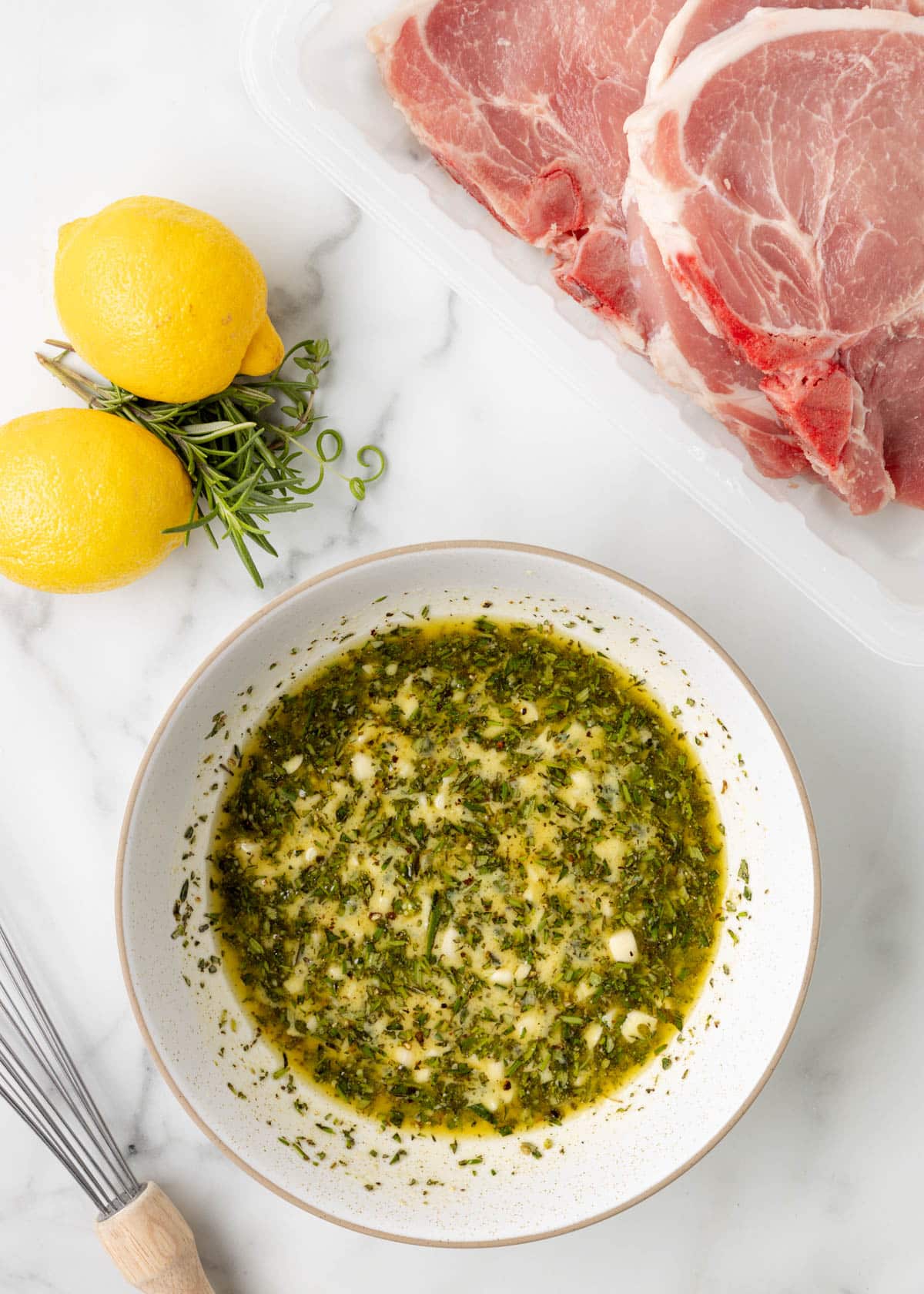 overhead of mixed lemon herb marinade in a white bowl next to whole lemons and a package of pork chops