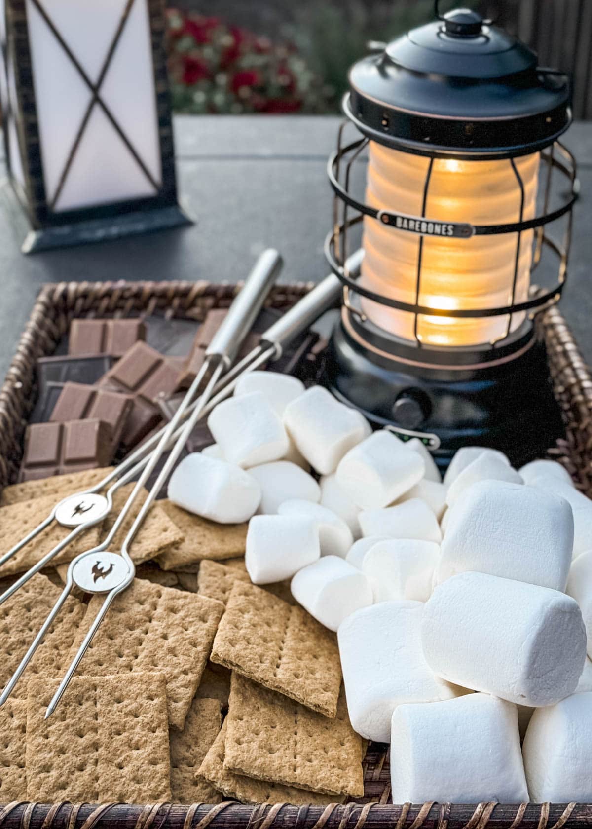 wicker tray filled with marshmallows, graham crackers, and chocolate with a lit lantern and marshmallow roasting sticks