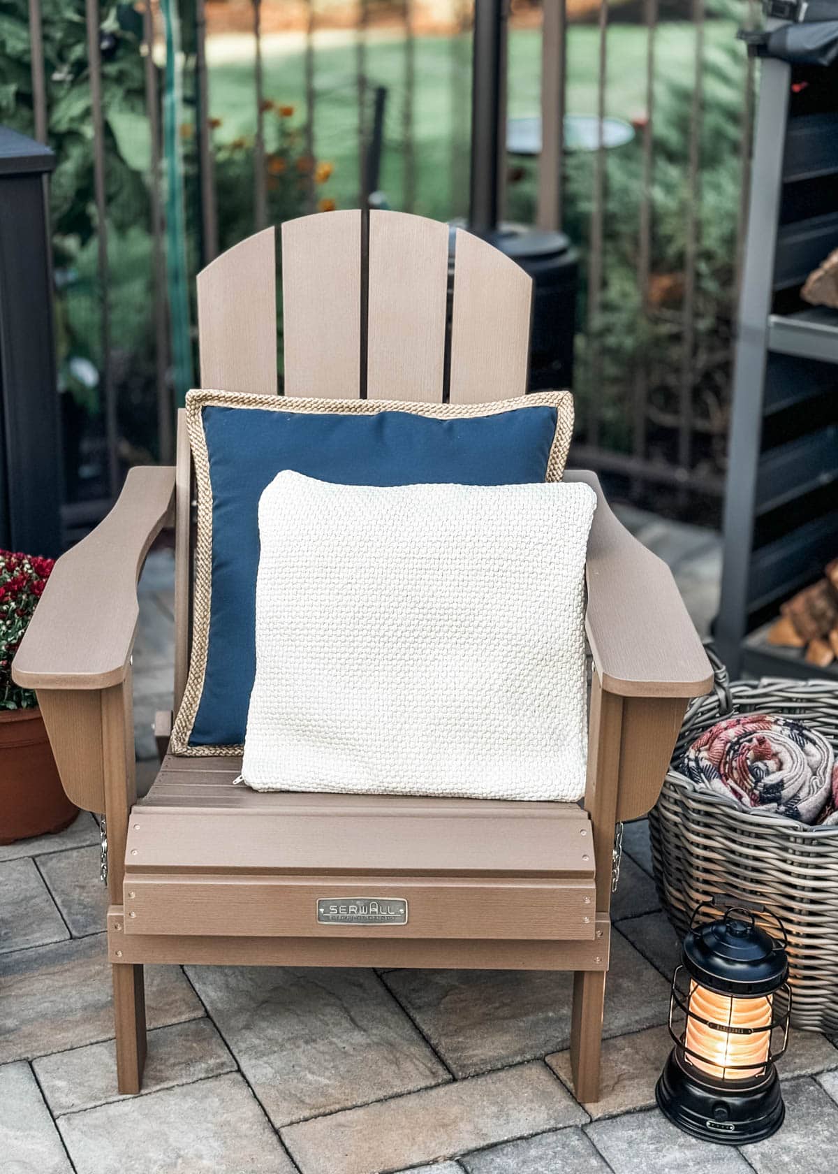 Adirondack chair with blue and ivory pillows on a patio next to a lantern, basket of blankets, and firewood rack