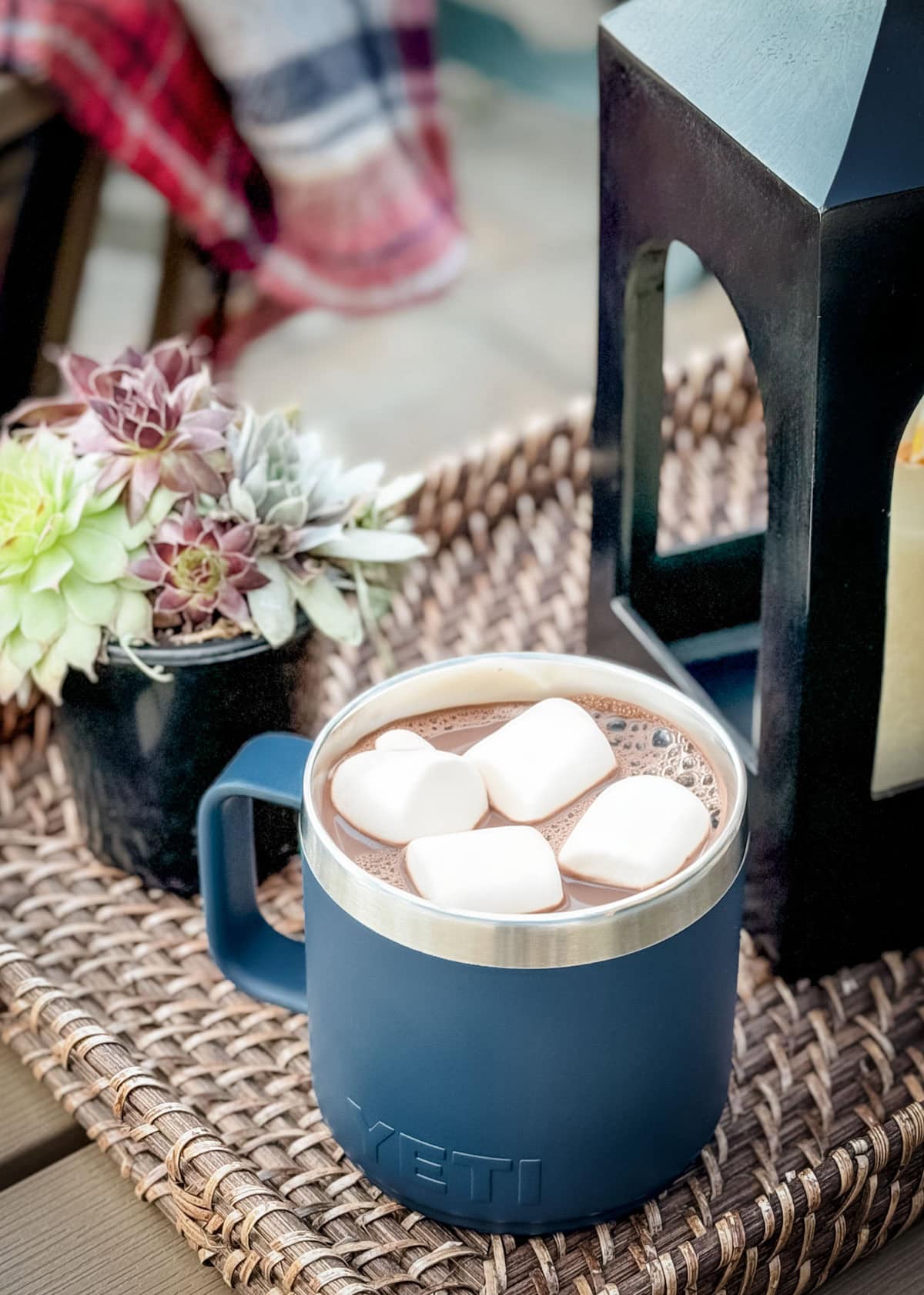 blue insulated mug filled with hot chocolate and marshmallows on a wicker tray with a black lantern and succulent plant