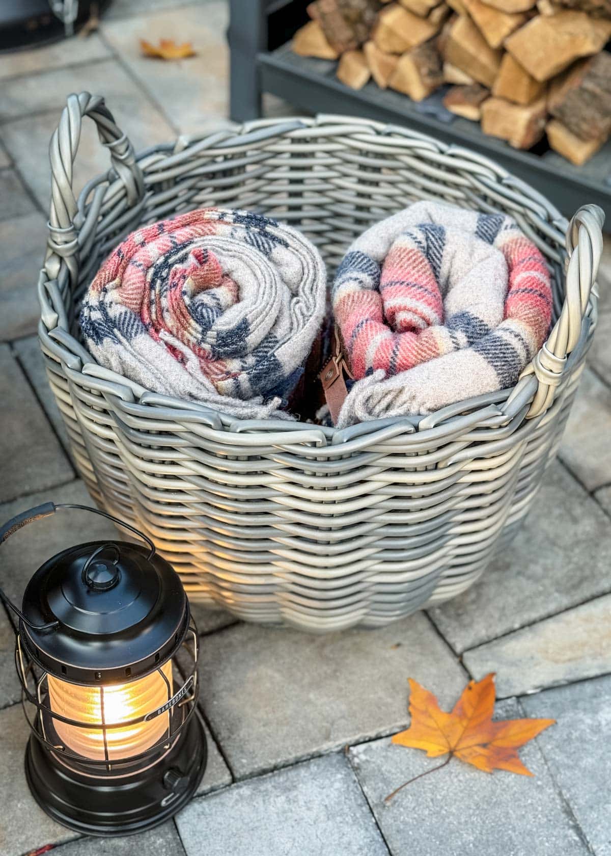 basket of wool blankets on a paver stone patio next to a lantern and a rack of firewood