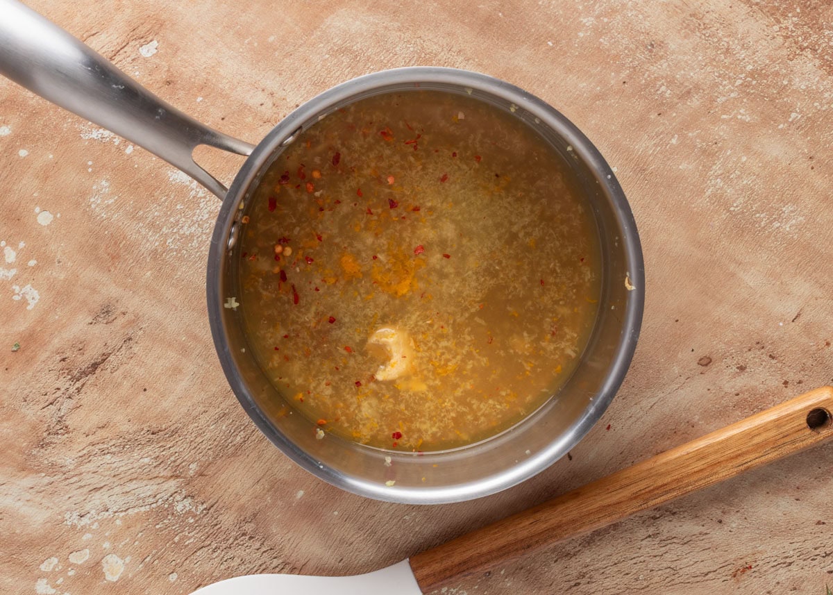 overhead of uncooked orange maple bourbon glaze in a stainless steel pot next to a white and wood handled rubber spatula
