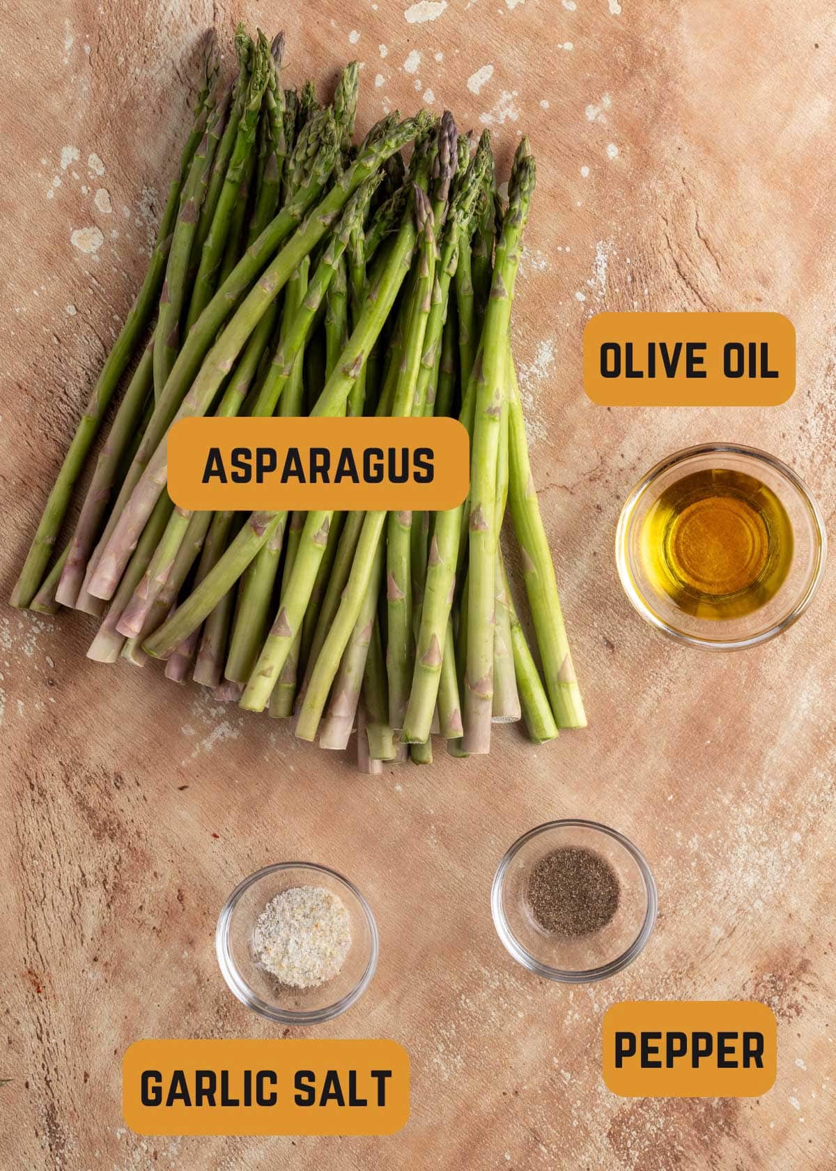 pile of raw asparagus next to bowls of olive oil, garlic salt, and pepper with text label overlays