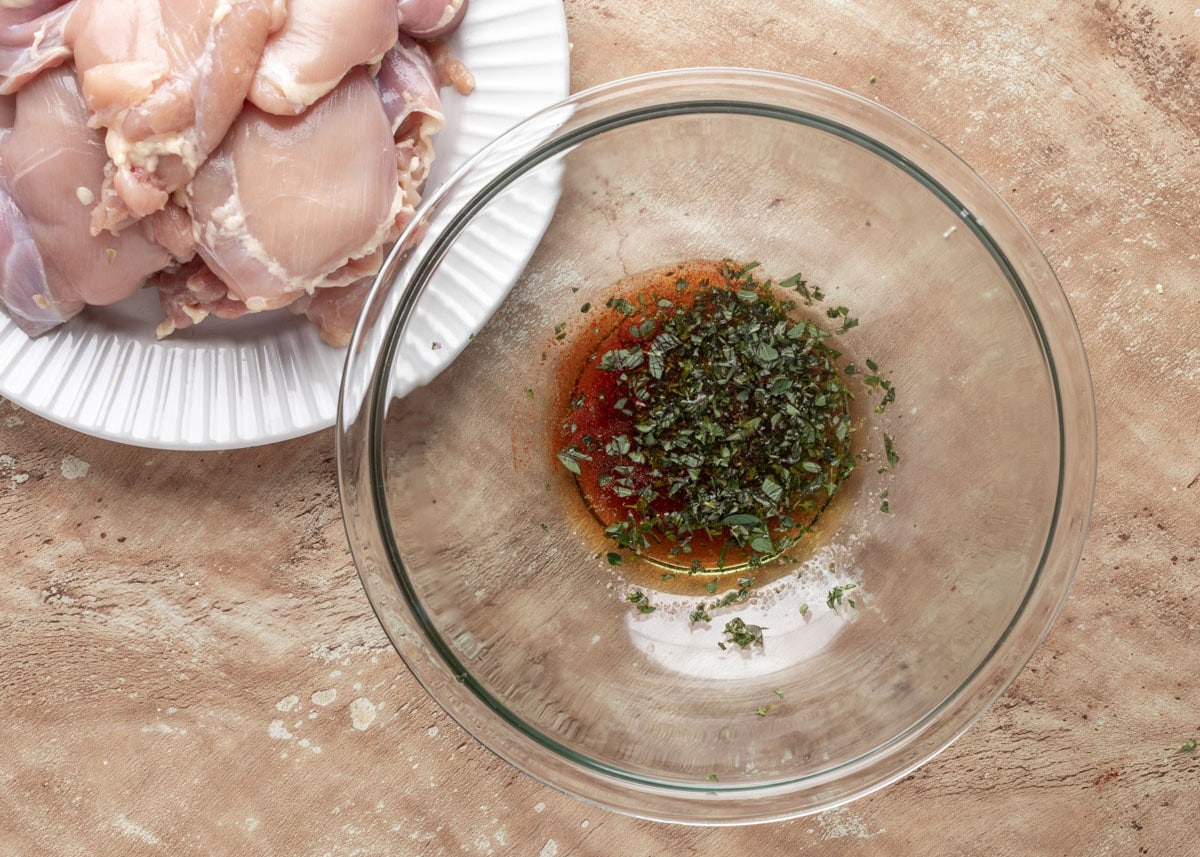 unmixed chicken marinade in a glass bowl next to raw chicken thighs on a white plate