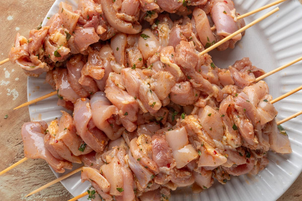 raw chicken souvlaki on bamboo skewers stacked on a white plate.