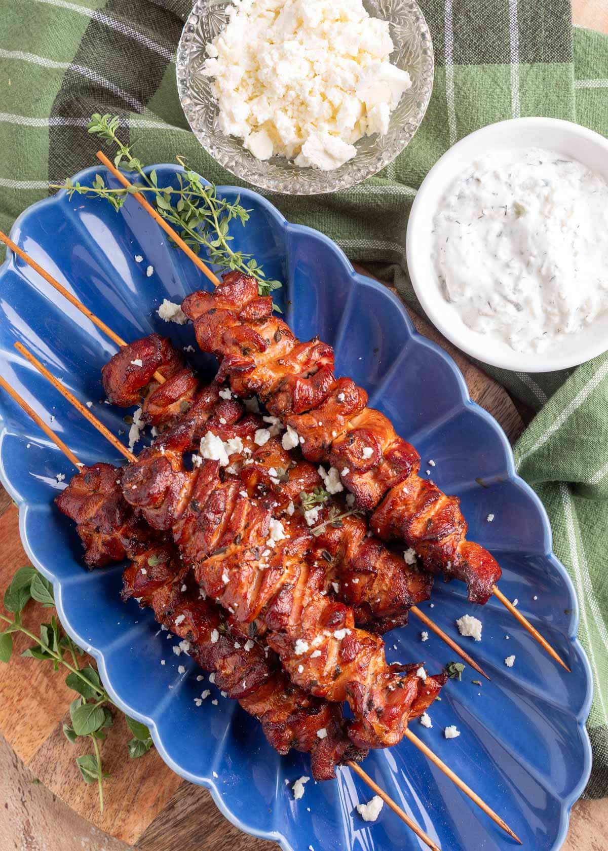overhead of smoked chicken souvlaki skewers on a blue scallpped platter next to bowls of feta and tzatziki witih a green and white striped napkin