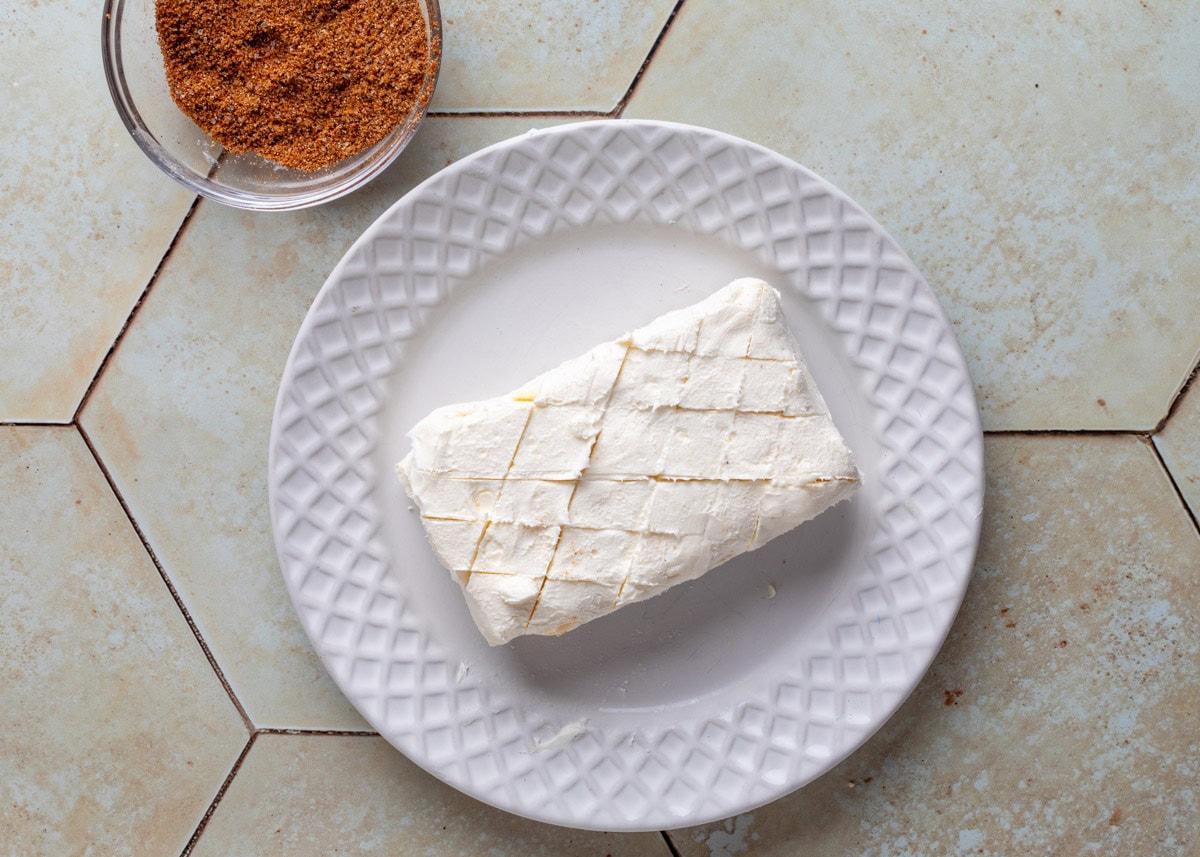 a scored block of cream cheese on a white plate next to a small bowl of spice rub