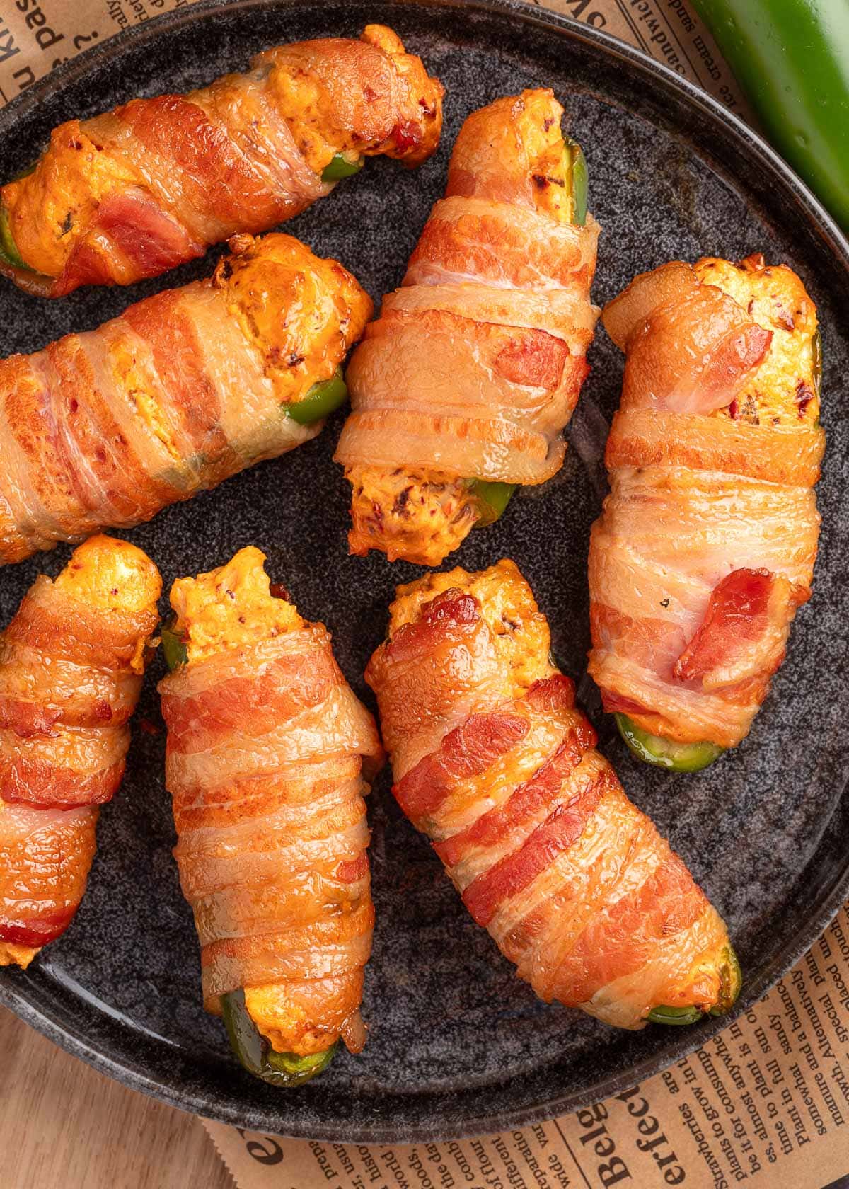 overhead of bacon wrapped smoked jalapeno poppers on a dark gray plate on top of a piece of beige newsprint