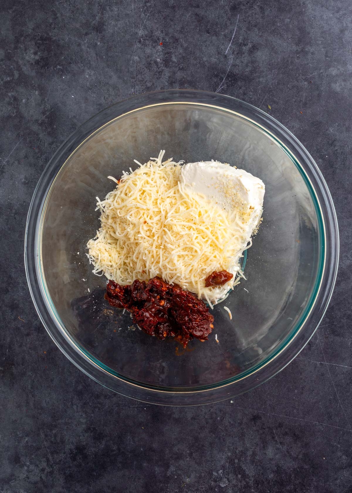 overhead of cream cheese, shredded gouda, and chopped chipotle in adobo in a glass bowl on a dark gray surface