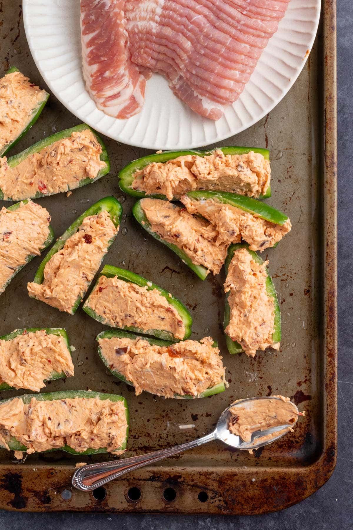 uncooked jalapeno popper halves stuffed with gouda and chipotle filling on a baking sheet next to a plate of raw bacon