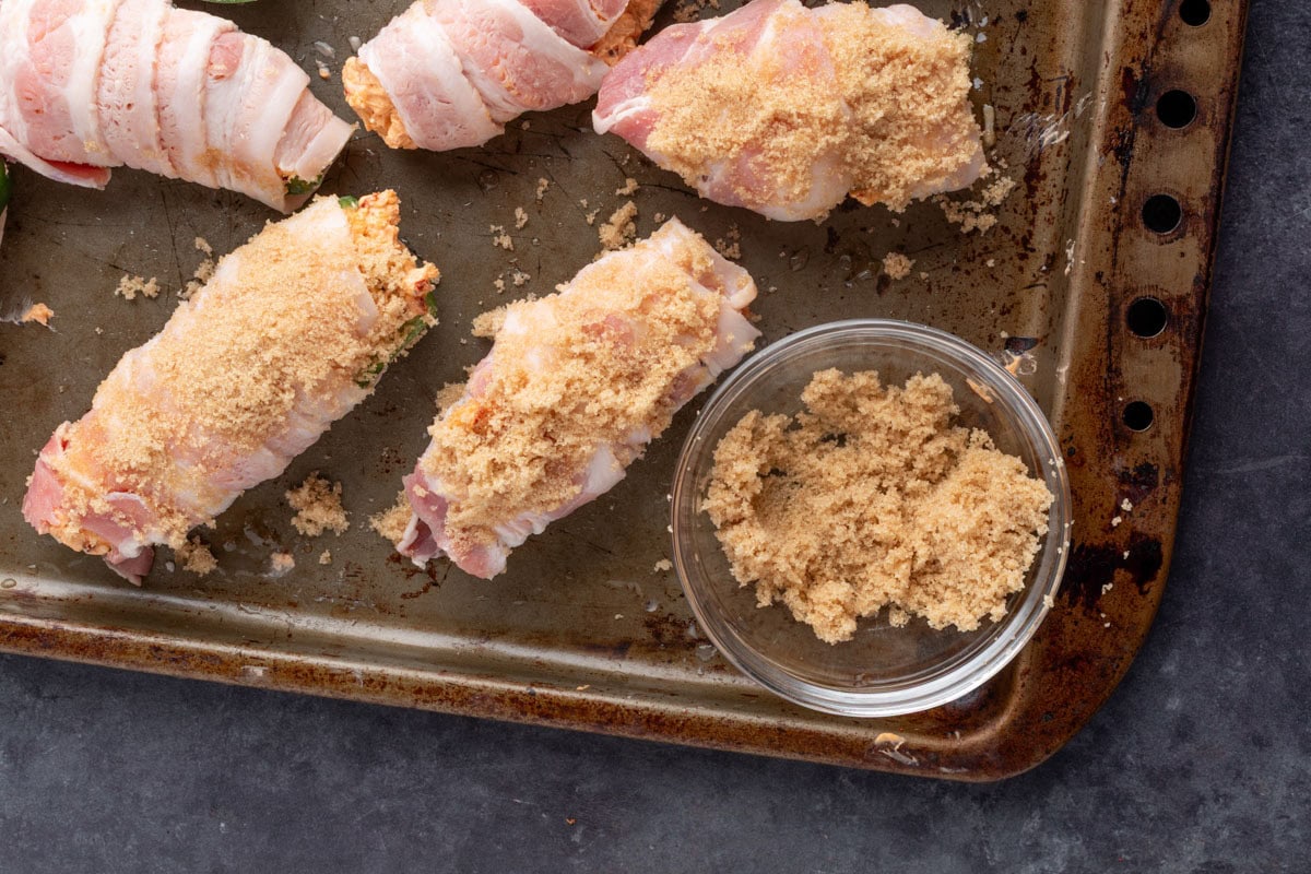 raw bacon wrapped jalapeno poppers on a baking sheet sprinkled with brown sugar