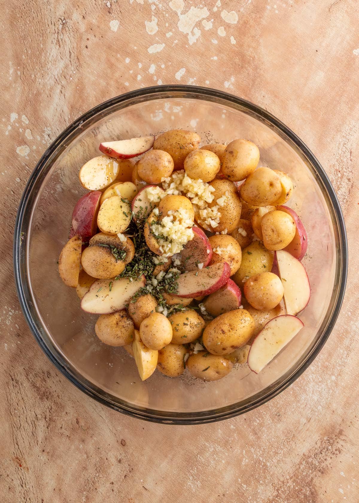 halved baby potatoes in a glass bowl with olive oil, garlic, and rosemary.