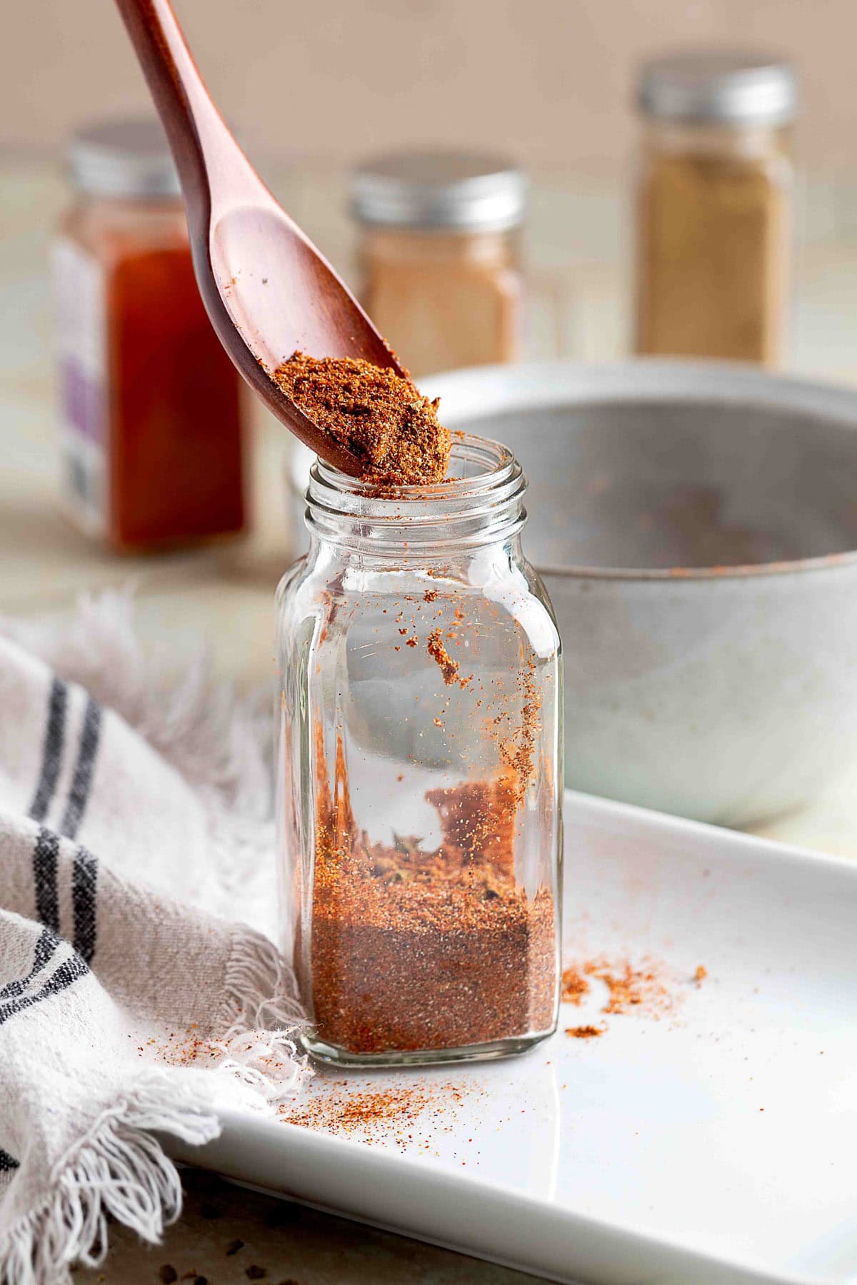 homemade fajita seasoning being poured into a glass spice jar with a wooden spoon on a white tray