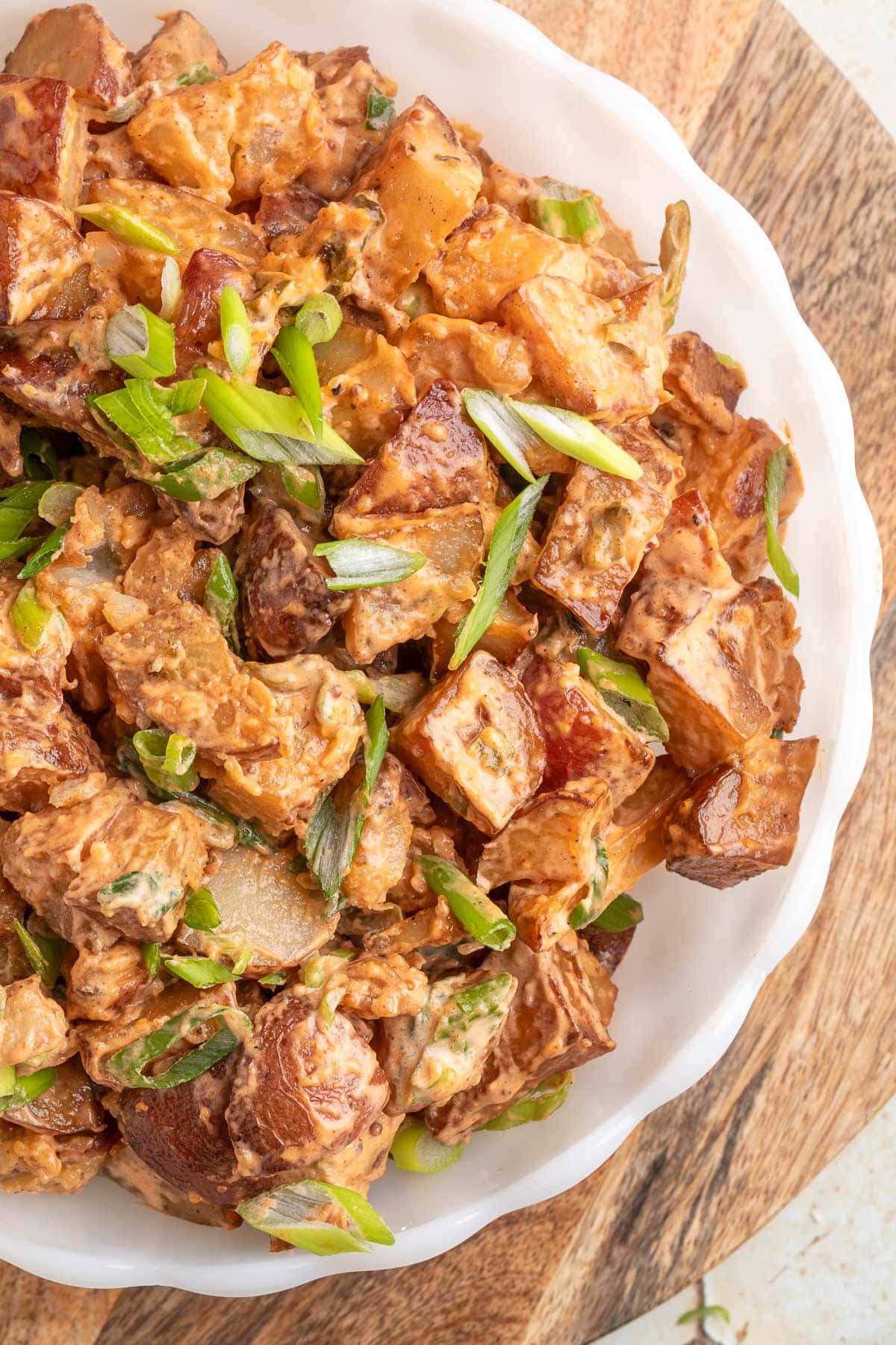smoked bbq potato salad garnished with scallions in a white scalloped bowl on a wood serving board