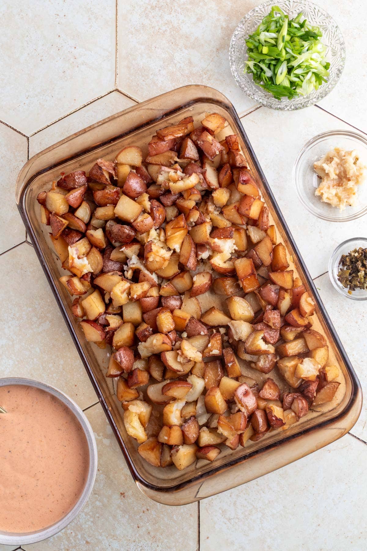 overhead of a pan of smoked potatoes, a bowl of potato salad dressing, and bowls of sliced scallions, chopped smoked garlic, and diced smoked jalapeno on an ivory tile surface