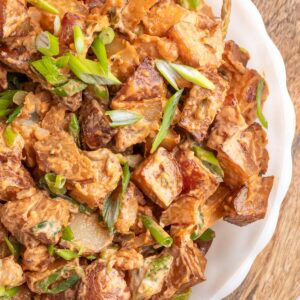 smoked bbq potato salad garnished with scallions in a white scalloped bowl on a wood serving board