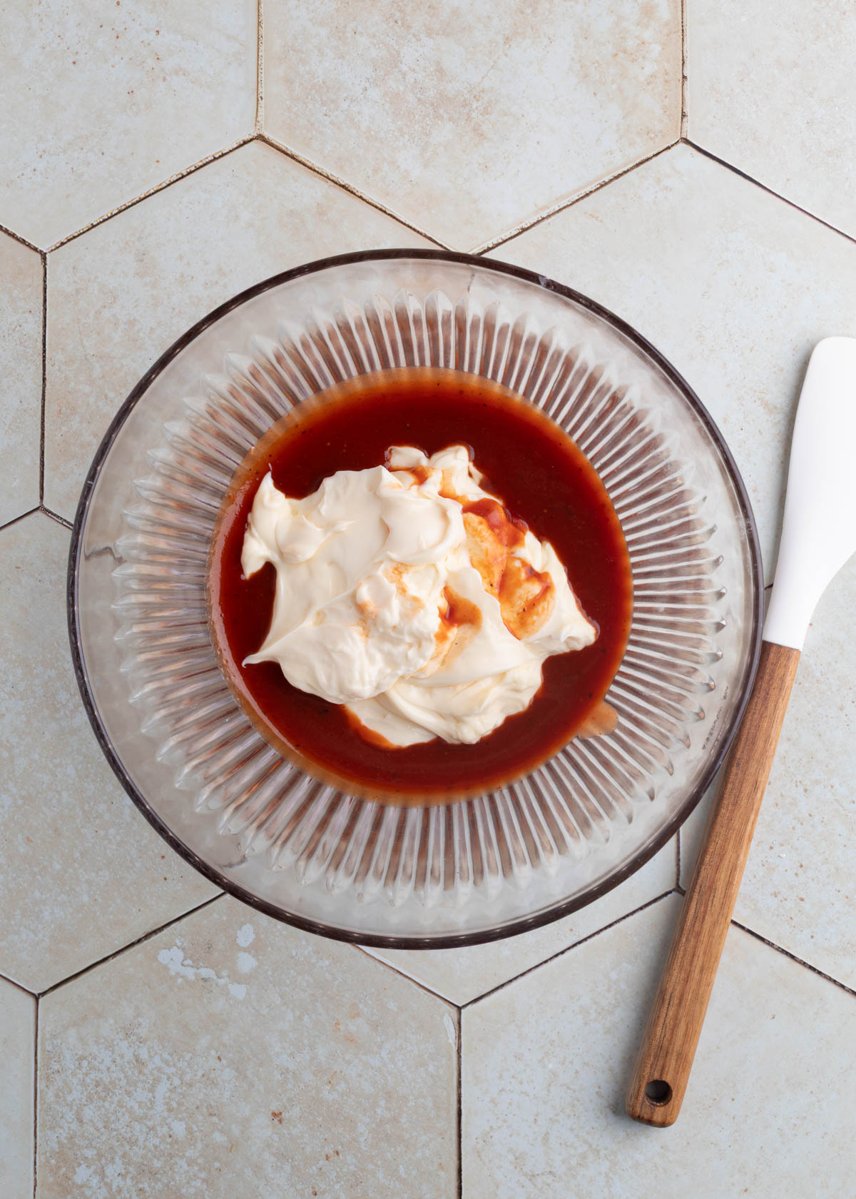 mayonnaise, bbq sauce, and grainy mustard in a glass bowl next to a white rubber spatula on an ivory tile surface